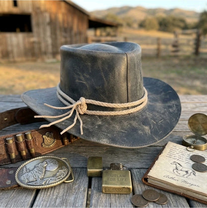 A distressed black leather cowboy hat with a beige rope band sits on a wooden table outdoors, styled with western accessories including a leather belt with brass buckle, a brass pocket watch, a small brass flask, an old book, and coins, with a blurred barn and hills in the background.