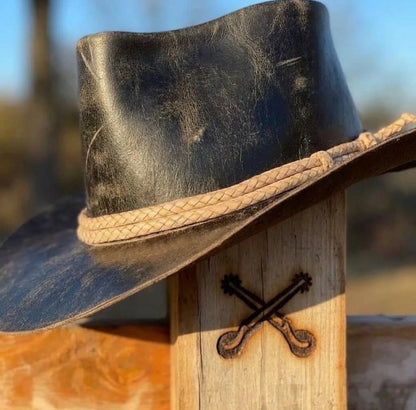 A black leather cowboy hat with a braided tan rope band, resting on a rustic wooden post outdoors.