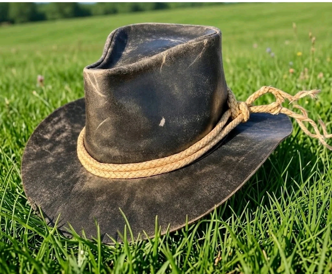 A dark brown distressed leather cowboy hat with a natural tan rope band, resting on a green grassy field.