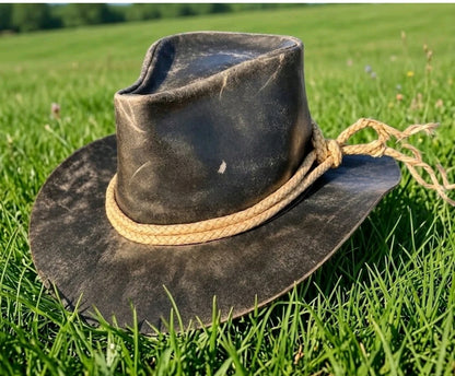 A dark brown distressed leather cowboy hat with a natural tan rope band, resting on a green grassy field.