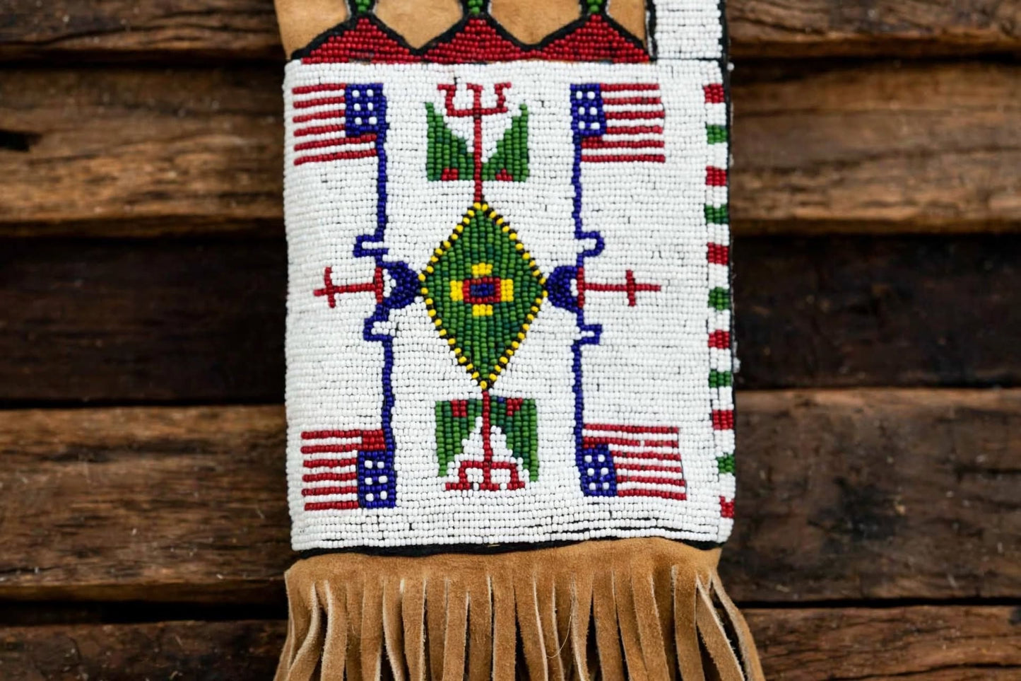A beaded fringed pouch or wall hanging with a Southwestern geometric design, featuring American flags, a green diamond motif, and tan fringe, displayed against a rustic wooden background.