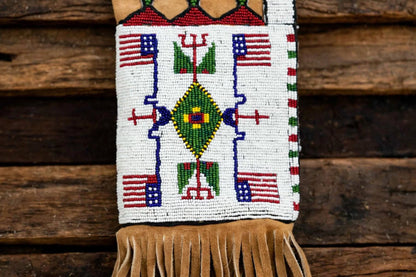 A beaded fringed pouch or wall hanging with a Southwestern geometric design, featuring American flags, a green diamond motif, and tan fringe, displayed against a rustic wooden background.