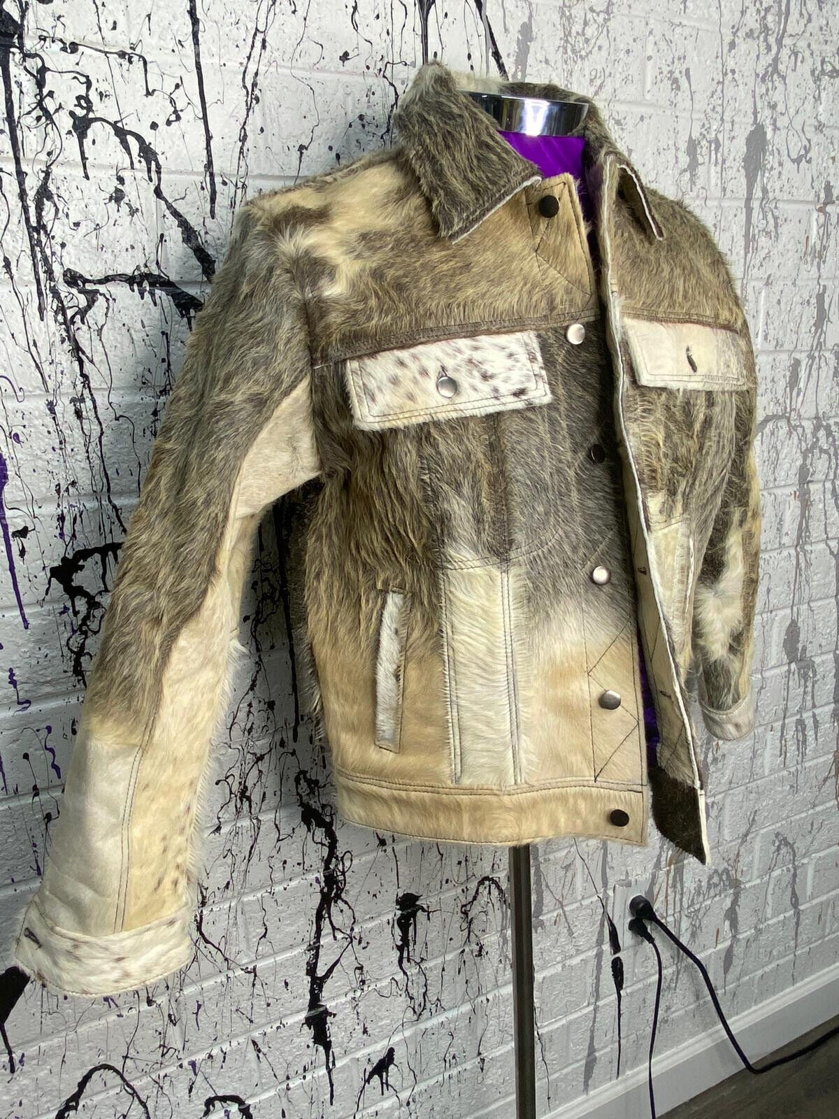 A men's cowhide jacket displayed on a mannequin against a textured background, featuring a natural brown and white cowhide pattern with patches of hair.