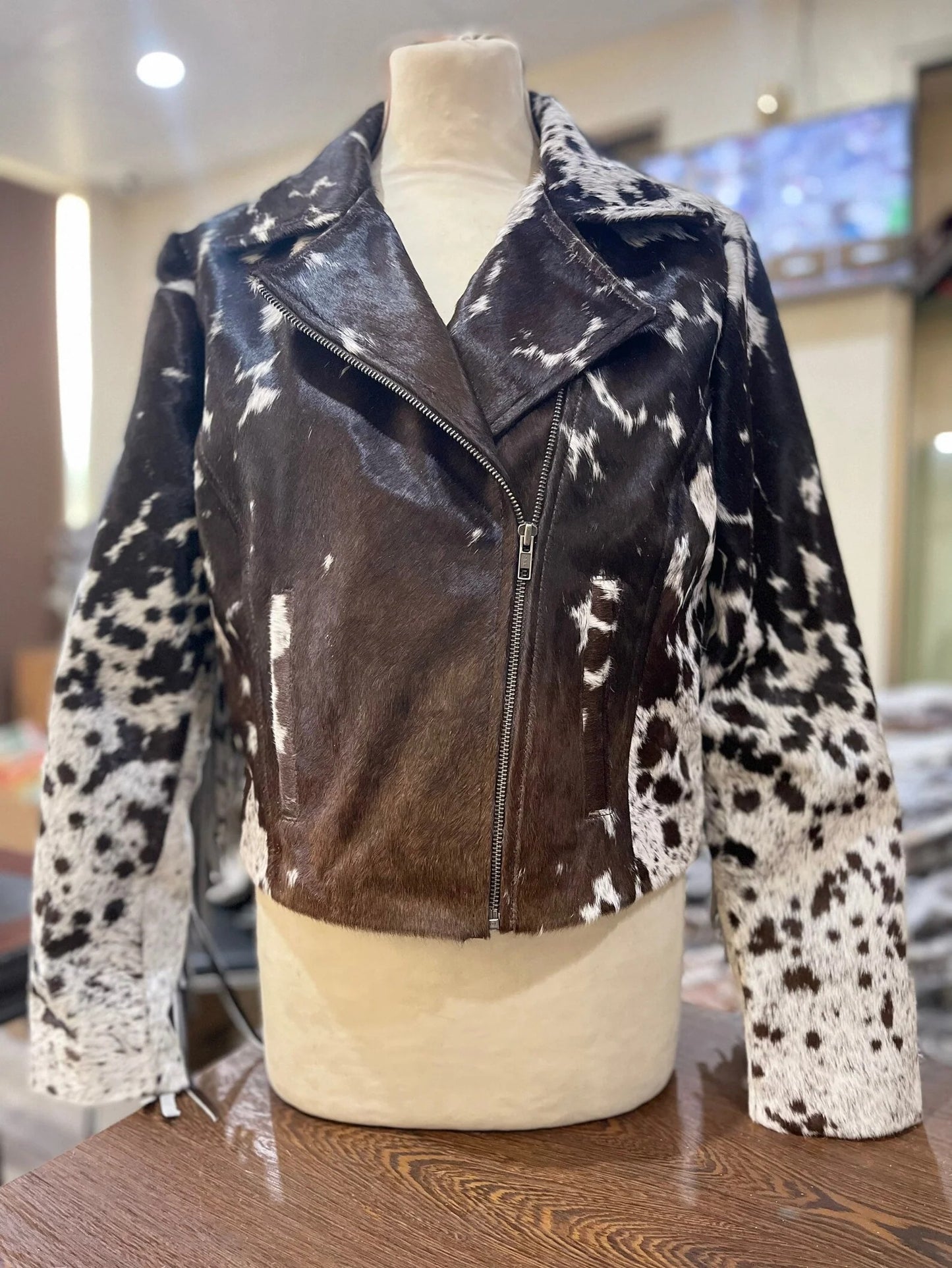 A cowhide jacket displayed on a mannequin, featuring a mix of black and white spots on the sleeves and a solid brown body with a zipper.