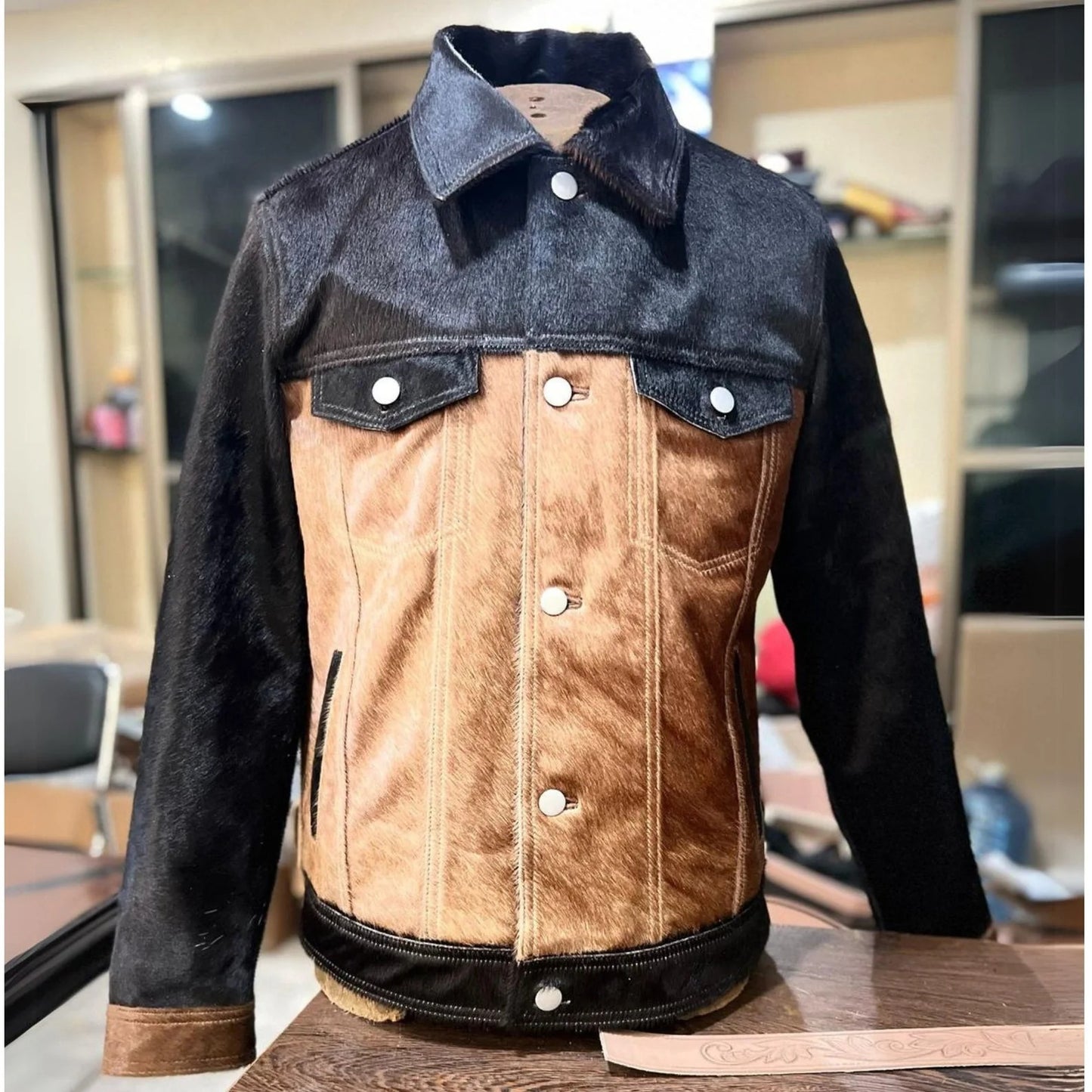 A men's cowhide jacket with a mix of brown and black colors, featuring a denim design, displayed on a table.