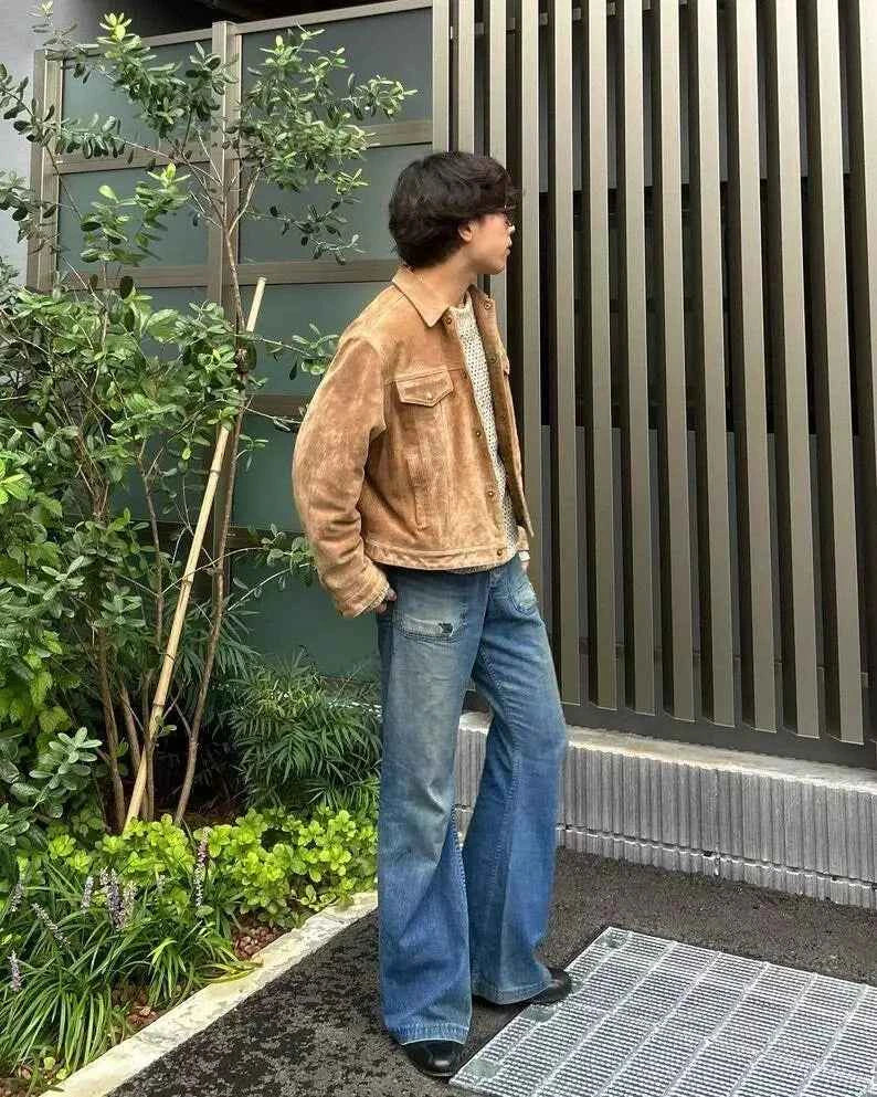 Person wearing a brown jacket and blue jeans standing outdoors next to a building with plants.