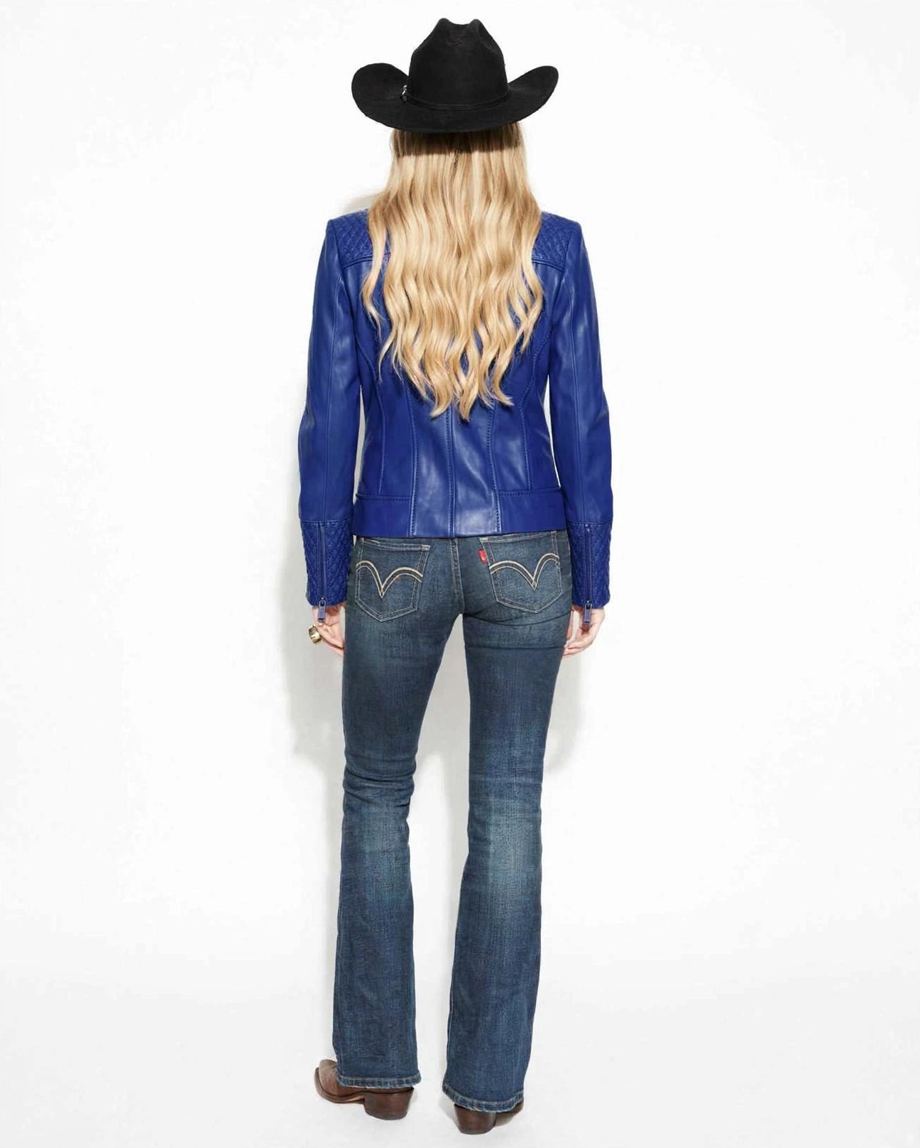 A woman shown from the back wearing a bright blue leather jacket, dark blue western flare jeans with embroidered details, brown boots, and a black cowboy hat against a plain white background.