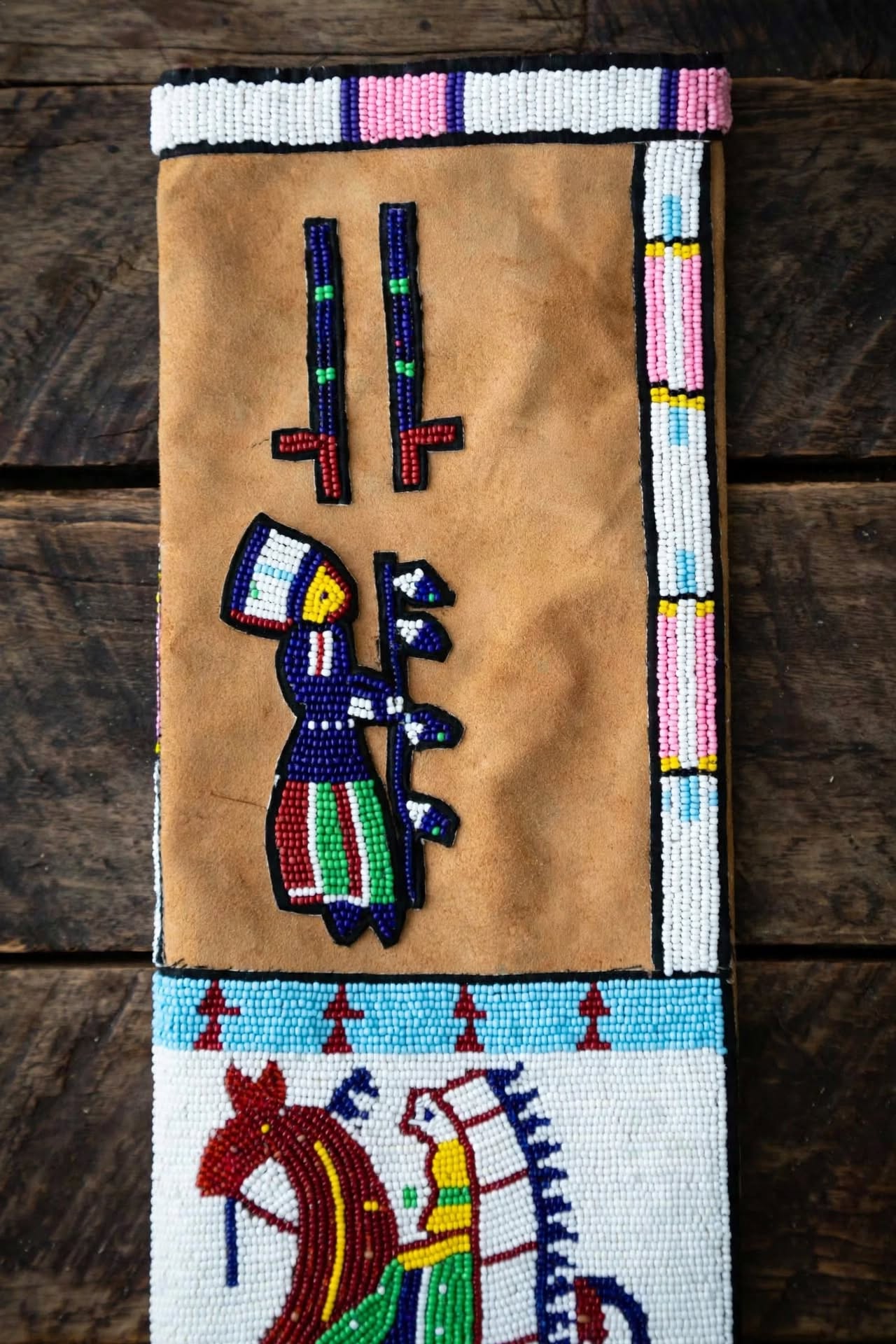 A rectangular tan leather or suede textile panel with beadwork, photographed flat on a dark wooden surface. The panel features multicolored bead-painted figures and geometric border patterns, including a central figure in blue, red, green, white, and yellow, and a horse motif near the bottom.