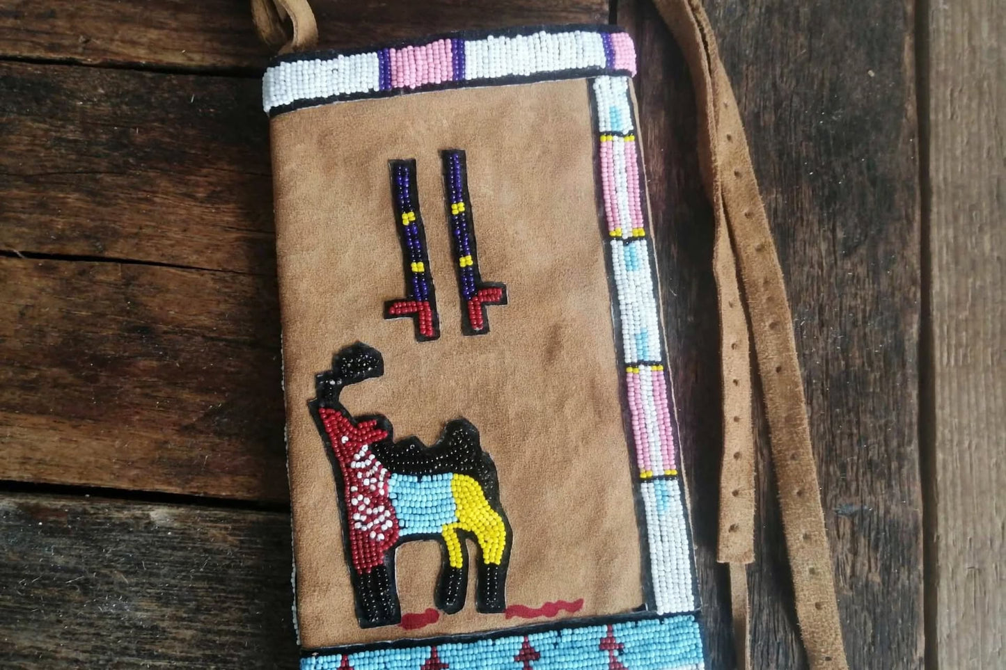 A tan suede crossbody wallet decorated with colorful beadwork, featuring a llama design and geometric borders, shown on a rustic wooden background.