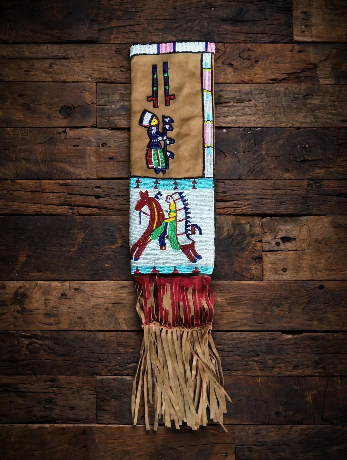 A handcrafted Native American style beaded fringed banner or wall hanging displayed against a rustic dark wood plank background. The textile features a tan top section with a colorful beaded figure, a white middle section with a red and yellow beaded horse motif, and a long fringe of tan leather and red tassels at the bottom.