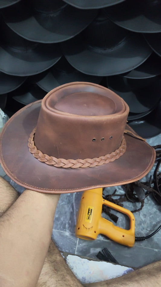 A brown leather cowboy hat with a braided band, held by a person's hand in front of a stack of black hats, with a yellow heat gun on a work surface below.