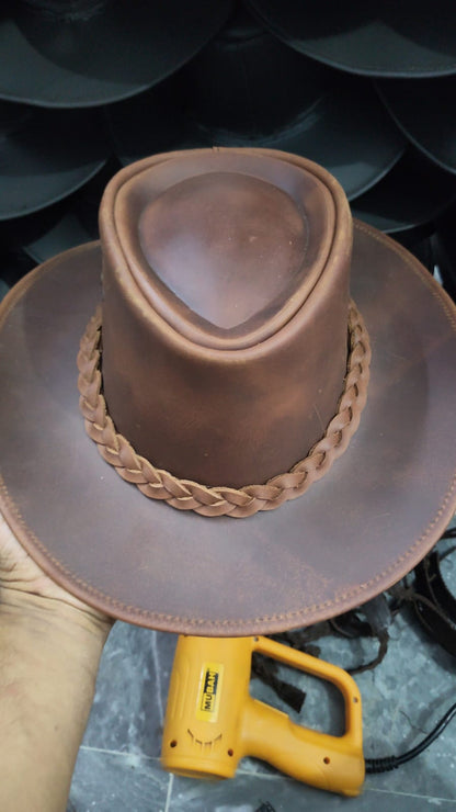 A brown leather cowboy hat with a wide brim and braided band, held by a hand. A yellow heat gun is visible below the brim, and other dark hats are stacked in the background.