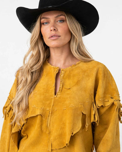 A woman wearing a black cowboy hat and a distressed mustard yellow suede fringe top, photographed against a clean white background.