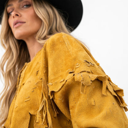 A close-up of a woman wearing a distressed mustard yellow suede fringe top and a black wide-brim hat against a plain white background.