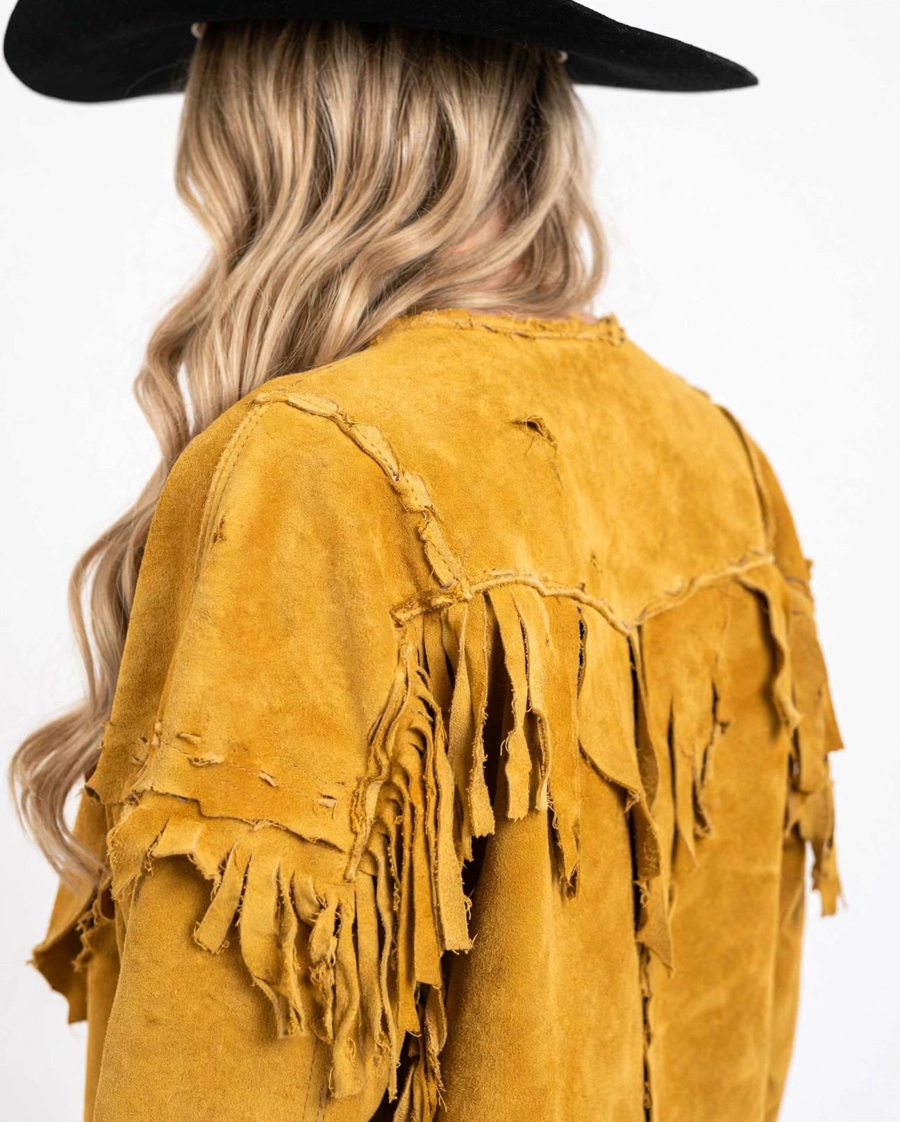 A close-up back view of a person wearing a distressed mustard yellow suede fringe jacket and a black wide-brim hat, against a plain white background.