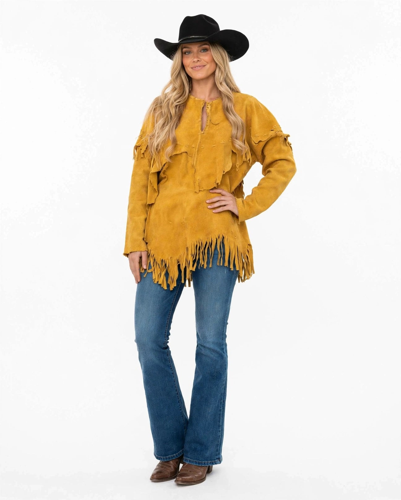 A woman modeling a mustard yellow suede fringe top with blue jeans, a black cowboy hat, and brown boots, photographed against a plain white studio background.